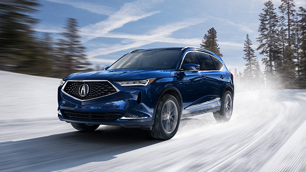 Front side-angled view of a blue Acura MDX driving a wintery road with trees and sky blurred in the background.	Vue latérale avant et en angle d’un Acura MDX bleu qui roule sur une route enneigée, avec le ciel et les arbres aux contours flous en arrière-plan.