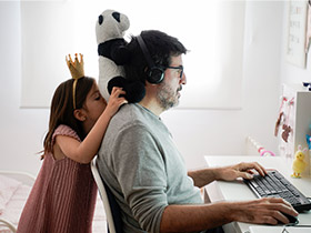 A man works at a computer while his daughter plays around behind him. / Un homme travaille devant un ordinateur pendant que sa fille joue derrière lui.